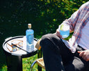 Man seated in a sunny garden holding an Azzurino cocktail with lime wheel, the Azzurino bottle and tortoiseshell sunglasses on a black side table.